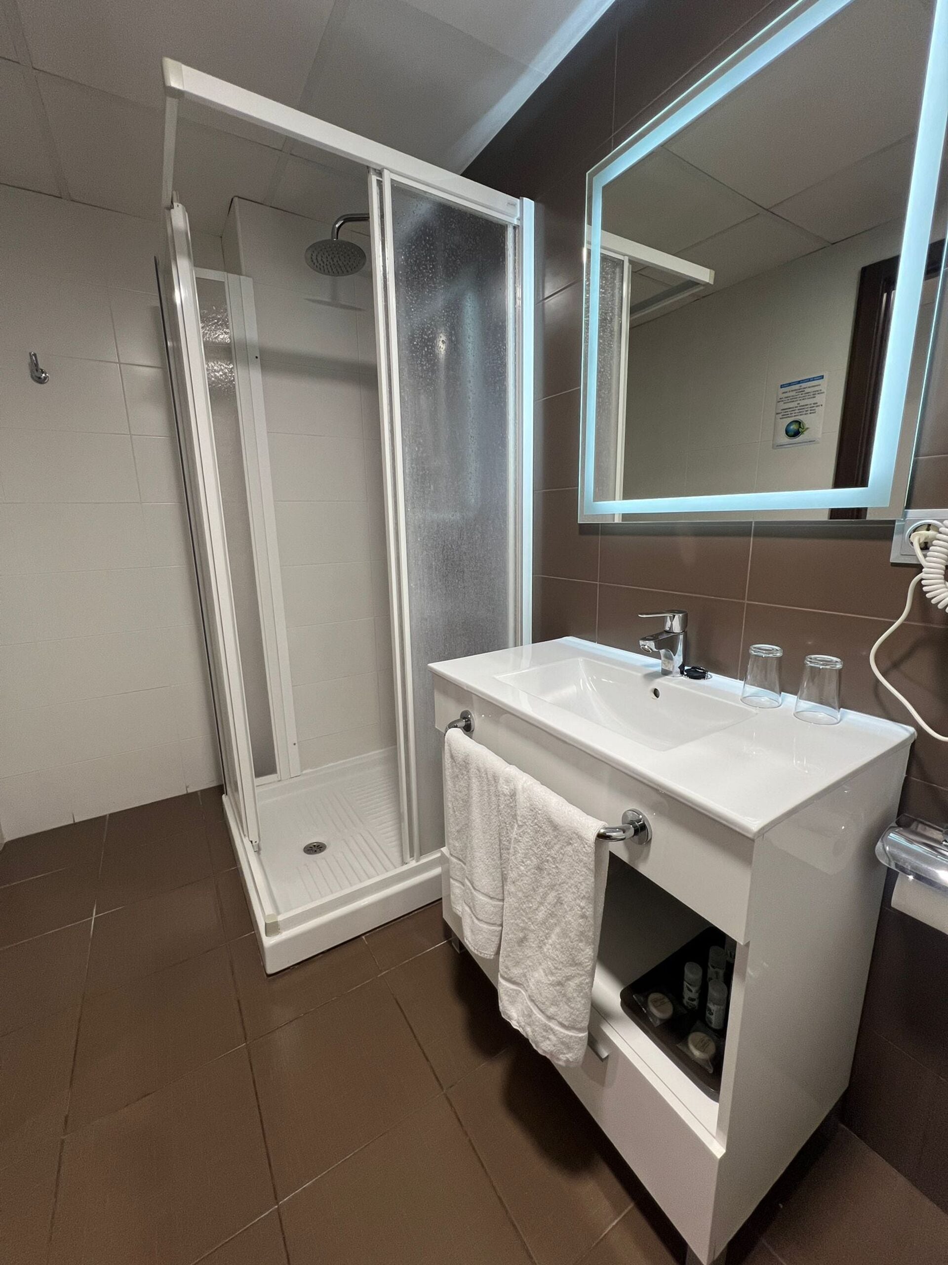 Un bagno moderno con doccia vetrata, un lavandino bianco con un asciugamano e uno specchio con cornice illuminata.