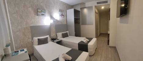 Habitación de hotel con dos camas, un escritorio pequeño, un televisor en la pared y un pasillo que conduce a otra habitación.