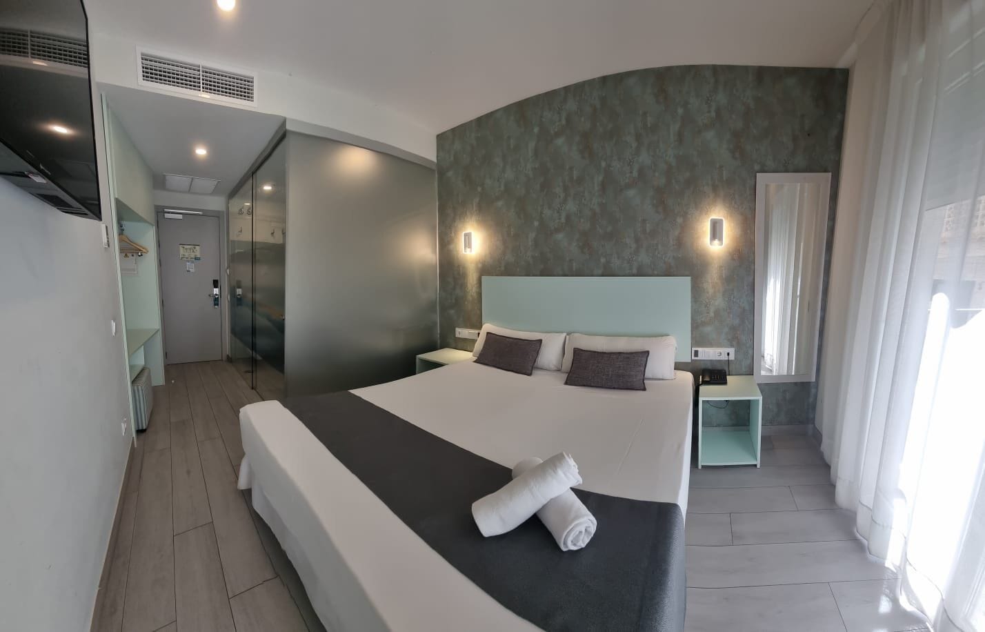 A modern hotel room with a large bed, a bedside table, a nightstand, a built-in wardrobe, and a bathroom with a glass shower enclosure.