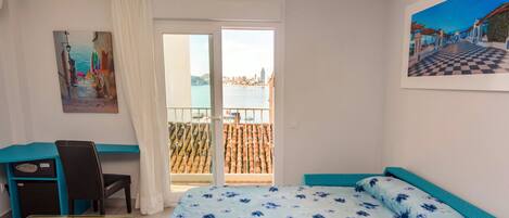 A bedroom with a bed, a desk, and a chair. There is a window with a view of a cityscape and a balcony with a railing.