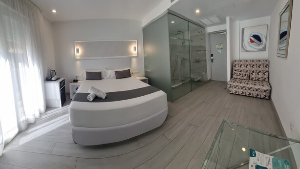 A modern hotel room with a curved bed, a glass-enclosed shower, a sofa, and a glass railing. - Hotel Clopy Rocamar (Benidorm)