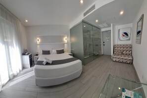 Habitación de hotel moderna con cama curvada, ducha con cerramiento de vidrio, sofá y barandilla de vidrio.