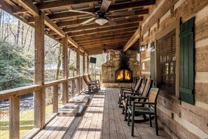 A wooden deck with a stone fireplace, wooden chairs, and a ceiling fan. - Indian Creek: Secluded, Beautiful Authentic Style Log Cabin in Gatlinburg (Cosby)