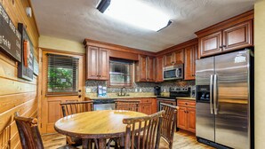 A kitchen with wooden cabinets, a stainless steel refrigerator, a wooden dining table with chairs, and a window with blinds. - Wildflower Haven: Beautiful Secluded Cabin in Gatlinburg, Just Minutes from Downtown Gatlinburg. (Sevierville)