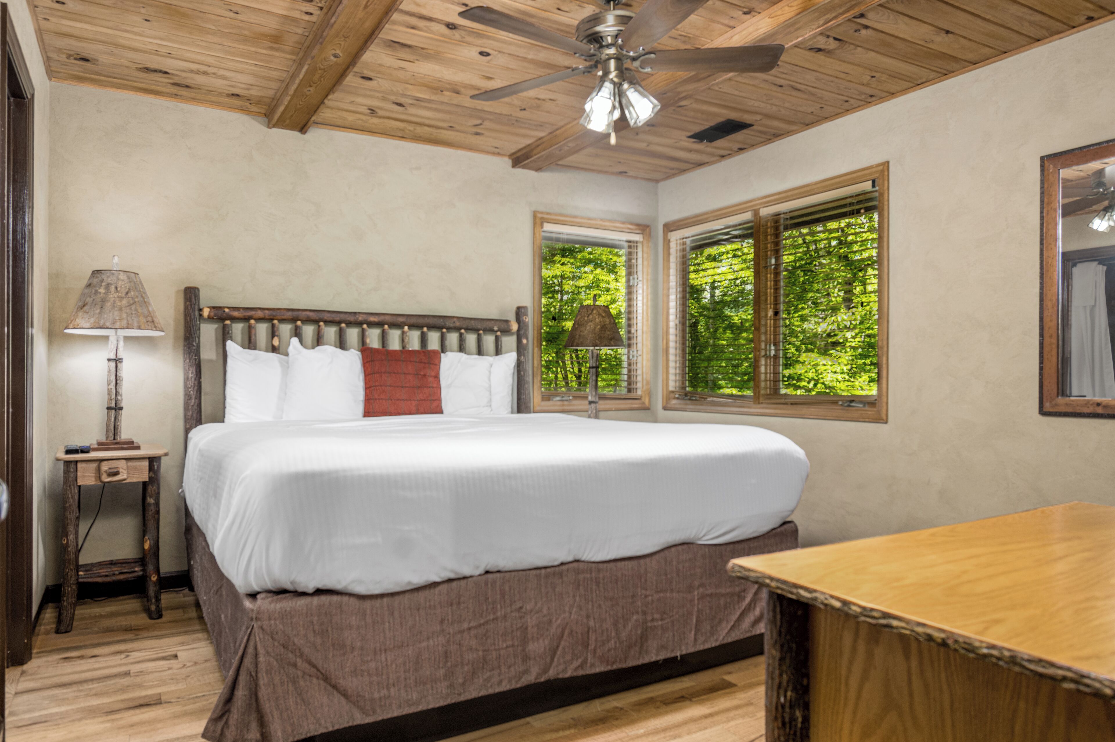 A bedroom with a wooden ceiling, a bed with white bedding, a nightstand with a lamp, and a window with a view of trees.