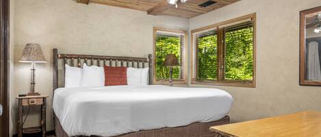 A bedroom with a wooden ceiling, a bed with white bedding, a nightstand with a lamp, and a window with a view of trees.
