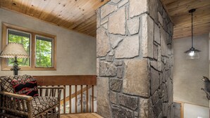 Ein Zimmer mit Kamin aus Stein, Holzboden und einem Schaukelstuhl mit gemusterten Kissen, sowie ein Fenster mit Blick auf Bäume.