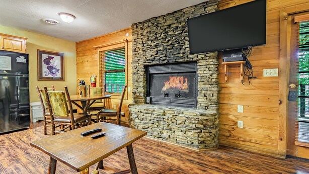 A wooden interior with a stone fireplace, a dining table, chairs, and a TV mounted on the wall. - Cub's Den: Beautifully Located, Cozy Cabin Surrounded by The Great Smoky Mountains (Gatlinburg)