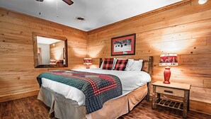 A wooden-paneled bedroom with a bed, a mirror, a framed picture, and a nightstand with a lamp. - Cub's Den: Beautifully Located, Cozy Cabin Surrounded by The Great Smoky Mountains (Gatlinburg)