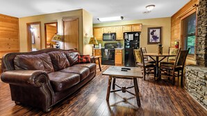 A cozy living room with a leather sofa, a wooden coffee table, and a dining area with chairs. A kitchen with wooden cabinets and appliances is visible in the background. - Cub's Den: Beautifully Located, Cozy Cabin Surrounded by The Great Smoky Mountains (Gatlinburg)