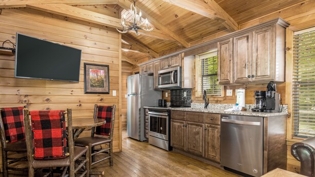 A wooden cabin interior with a kitchen featuring a refrigerator, microwave, stove, and dishwasher. There is a dining area with a table and chairs, and a television mounted on the wall.