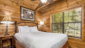 A wooden cabin bedroom with a bed, two bedside lamps, a ceiling fan, a framed picture on the wall, and a window with blinds.