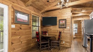 A wooden cabin interior with a dining table, chairs, a television, and a kitchen area.