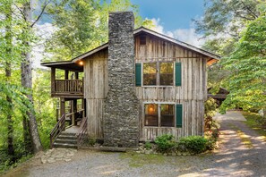 A rustic wooden cabin with a stone chimney, a wooden deck, and a gravel driveway surrounded by trees. - Valentines View: Majestic Lodge with Awe-Inspiring Views (Sevierville)