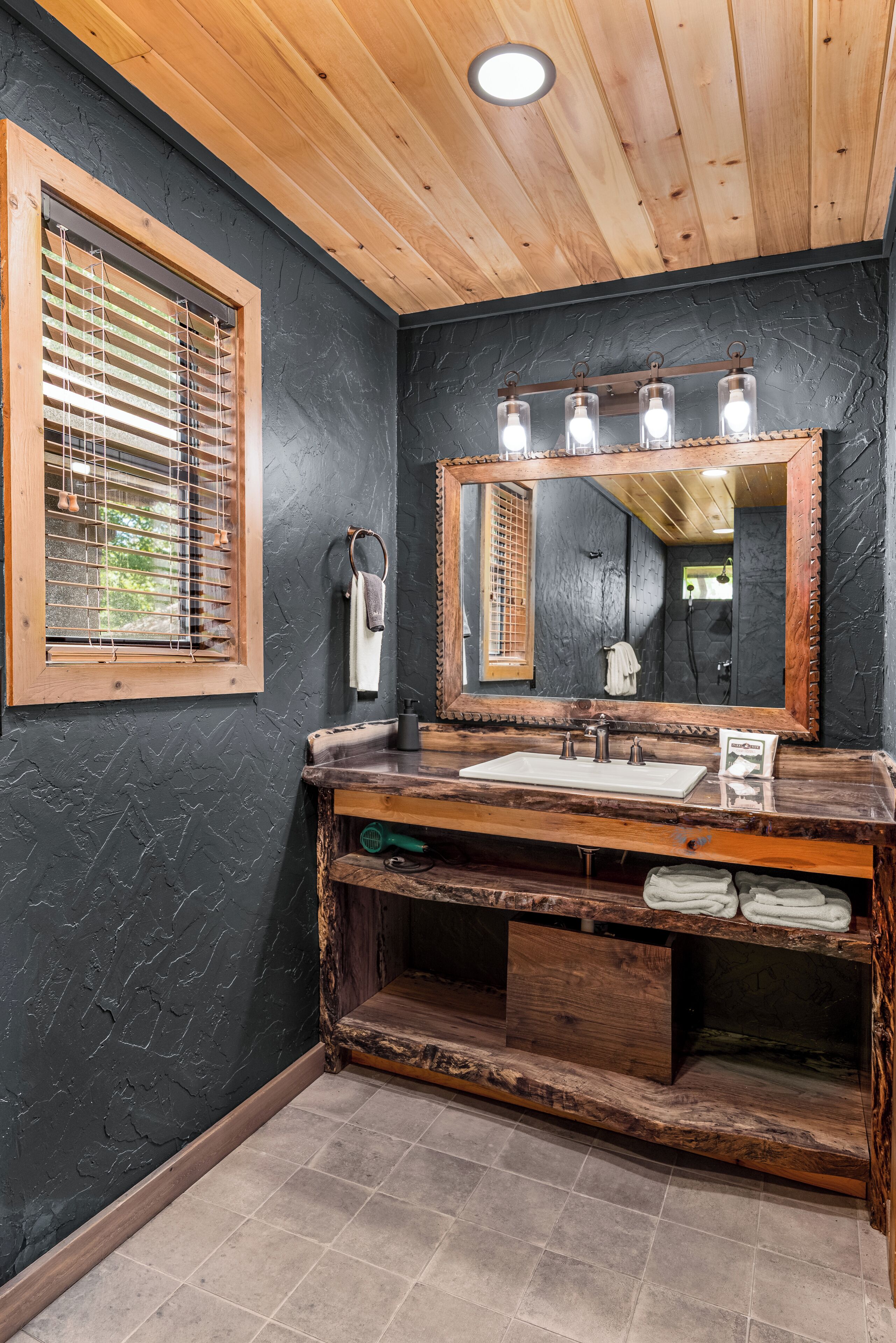 Une salle de bain avec un meuble-lavabo en bois, un grand miroir et une fenêtre avec des stores.