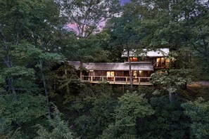 Une cabane entourée d’une forêt dense, dotée d’une terrasse en bois et de grandes fenêtres.