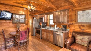 A rustic cabin-style kitchen with wooden walls, a dining area with a red and black checkered tablecloth, and a leather couch.
