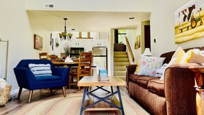 A living room with a blue armchair, a brown sofa, and a wooden coffee table. There is a kitchen in the background with white cabinets and a staircase leading to another level. - Charming, Farmhouse Chic, Cozy|Fireplace|12 Min to Town|Beautiful View (Galena)