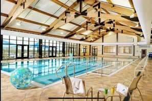 Une grande piscine intérieure dotée d’un haut plafond et de poutres en bois, entourée de grandes fenêtres et offrant une vue sur l’extérieur.