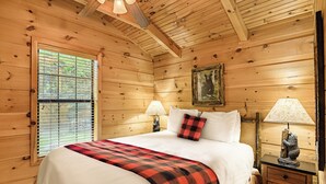 Un dormitorio de cabaña de madera con una cama, dos lámparas de noche, un ventanal con persianas y una estatua de oso sobre una mesita de noche.