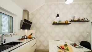 A modern kitchen with a built-in oven, a sink, and a countertop with a cutting board, knife, and bottle. There are two hanging pendant lights and a window with blinds.