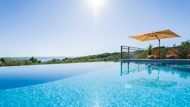 An infinity pool with clear blue water, a wooden deck with lounge chairs and an umbrella, and a view of the sea and hills in the background.