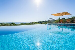 An infinity pool with clear blue water, a wooden deck with lounge chairs and an umbrella, and a view of the sea and hills in the background.
