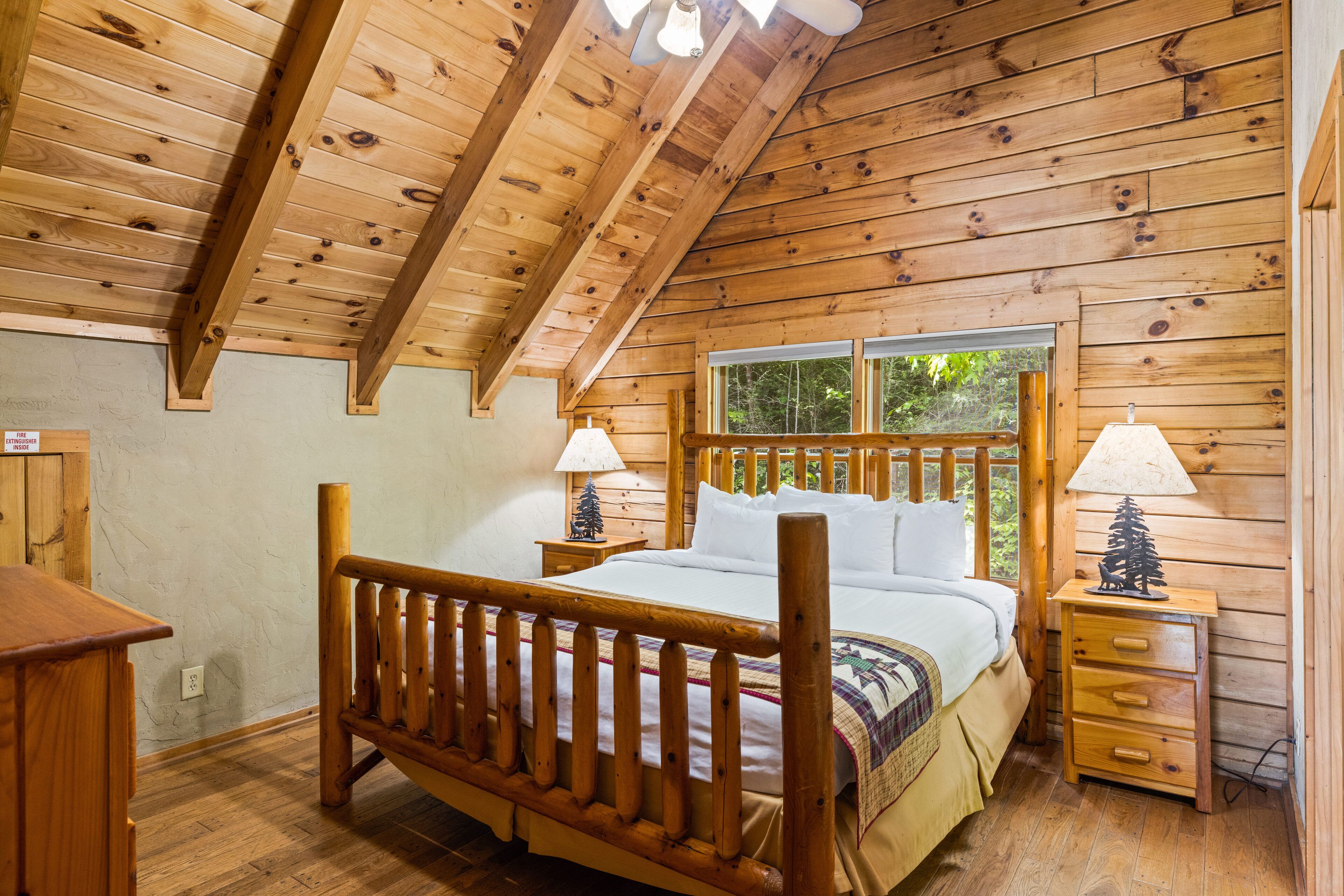 Un dormitorio de cabaña de madera con una cama, dos mesitas de noche, un aparador y una ventana con vistas a árboles.