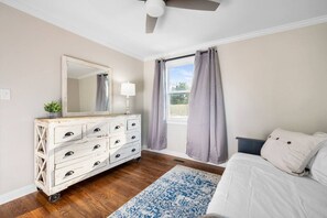 Un dormitorio con cama, cómoda, espejo, lámpara, ventana con cortinas y ventilador de techo.