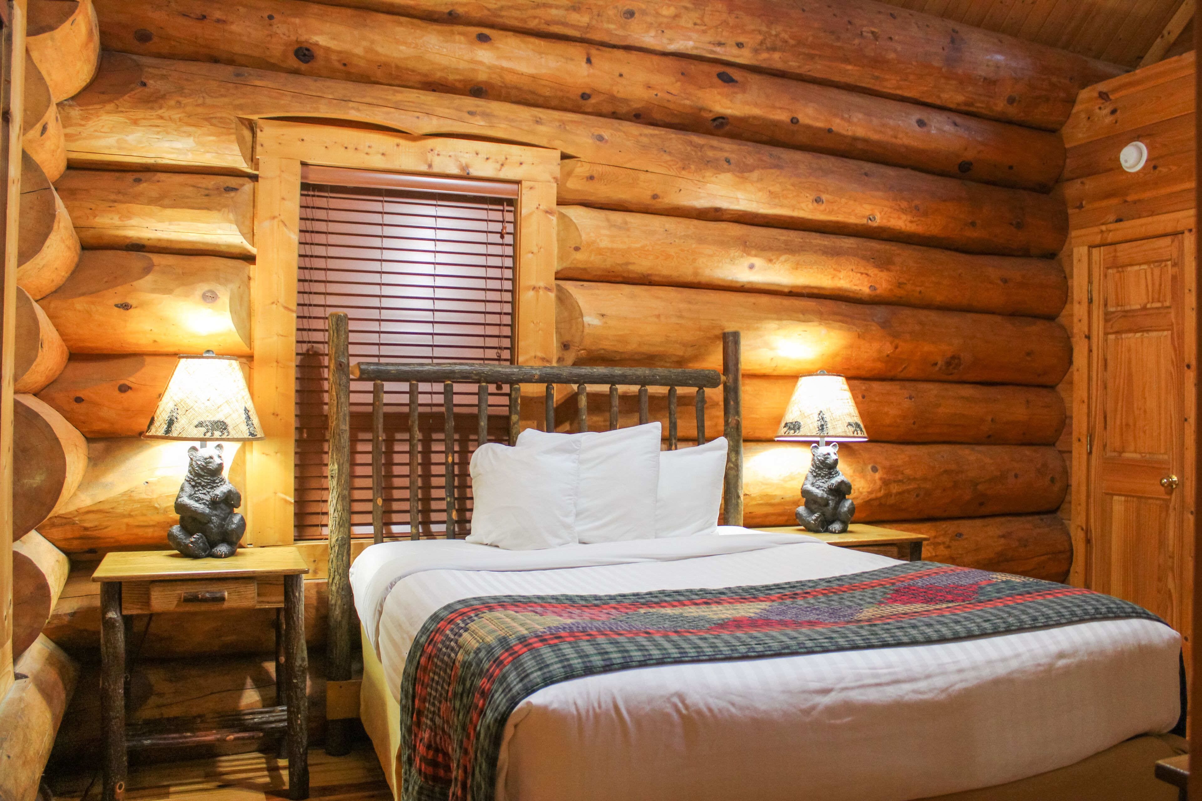 A log cabin-style room with a wooden bed, two nightstands with lamps, and a window with blinds.