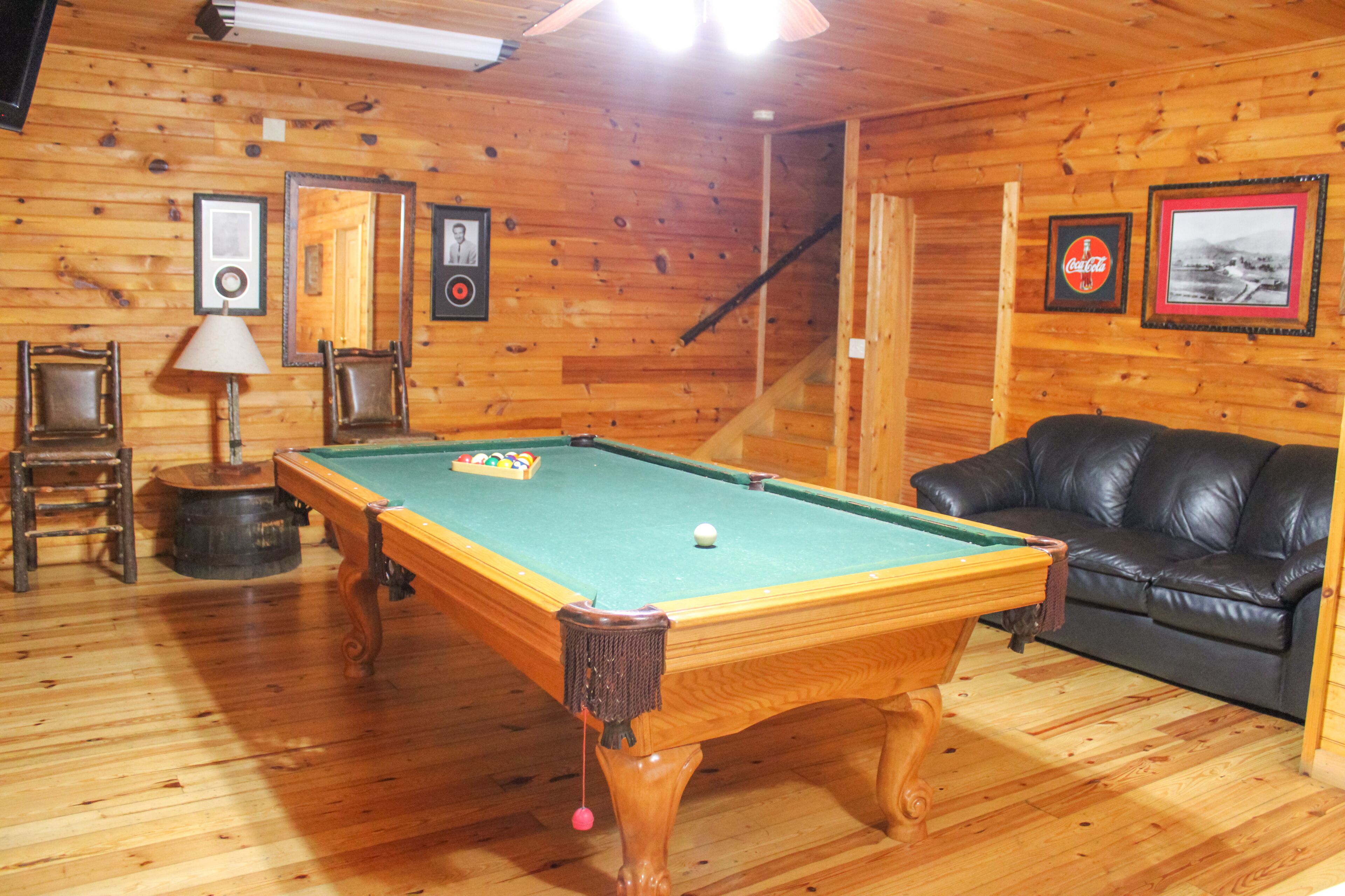 A pool table with a green felt surface, a black leather sofa, wooden walls, and framed pictures on the wall.