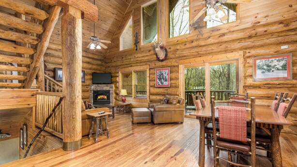 A log cabin interior with a fireplace, wooden furniture, and large windows.