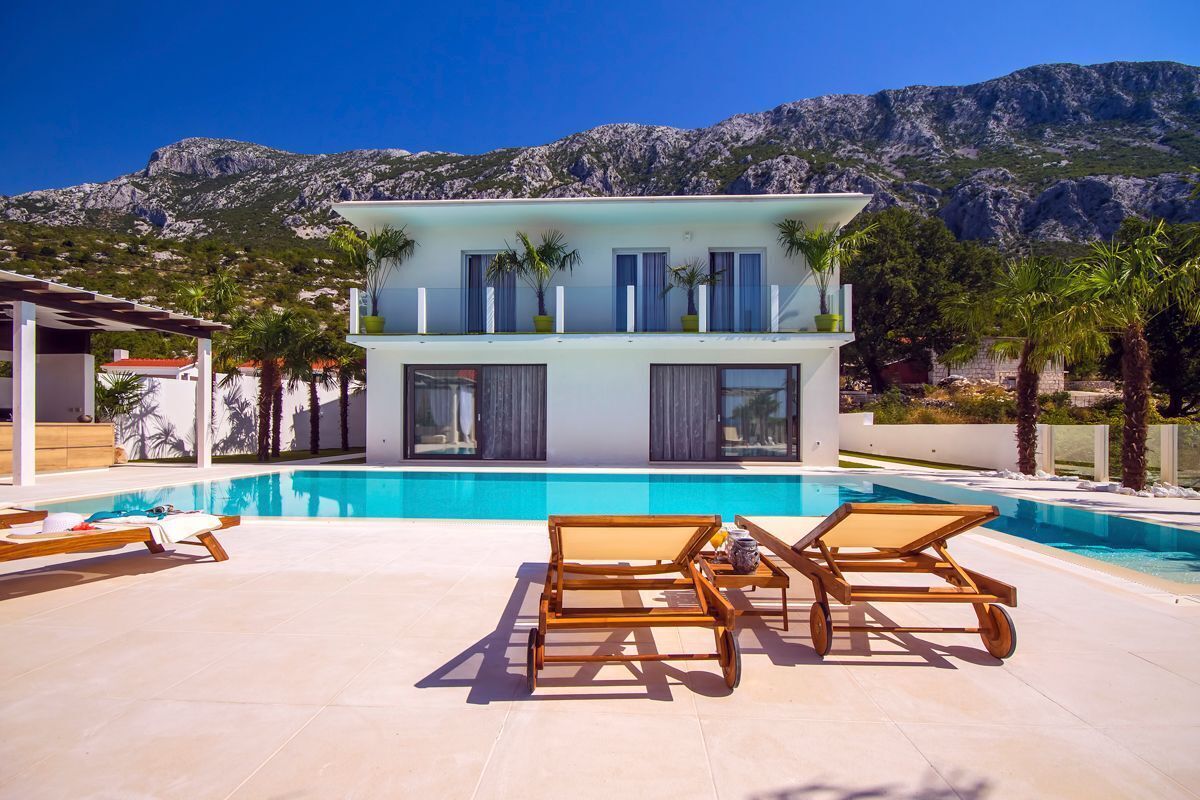 A modern villa with a pool, two lounge chairs, and a mountain backdrop.