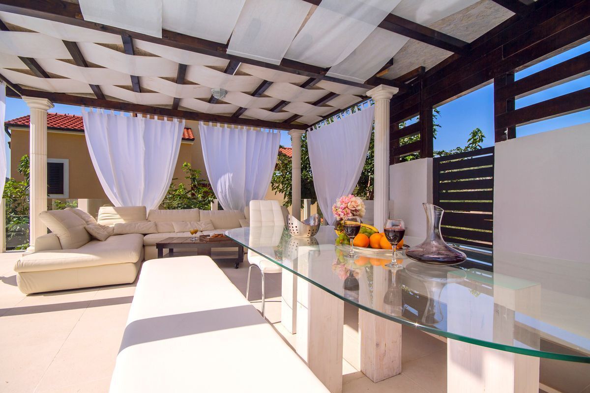 A modern outdoor living area with a glass table, beige sofa, and white curtains.