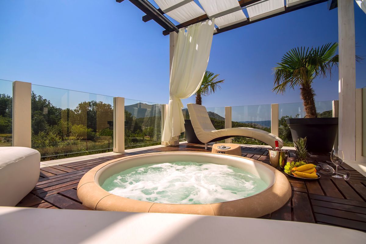 A hot tub on a wooden deck with a glass balustrade, a chair, and a table with fruits and a bottle.