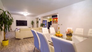 A modern living room with a beige sofa, a dining table with white chairs, and a TV mounted on the wall.