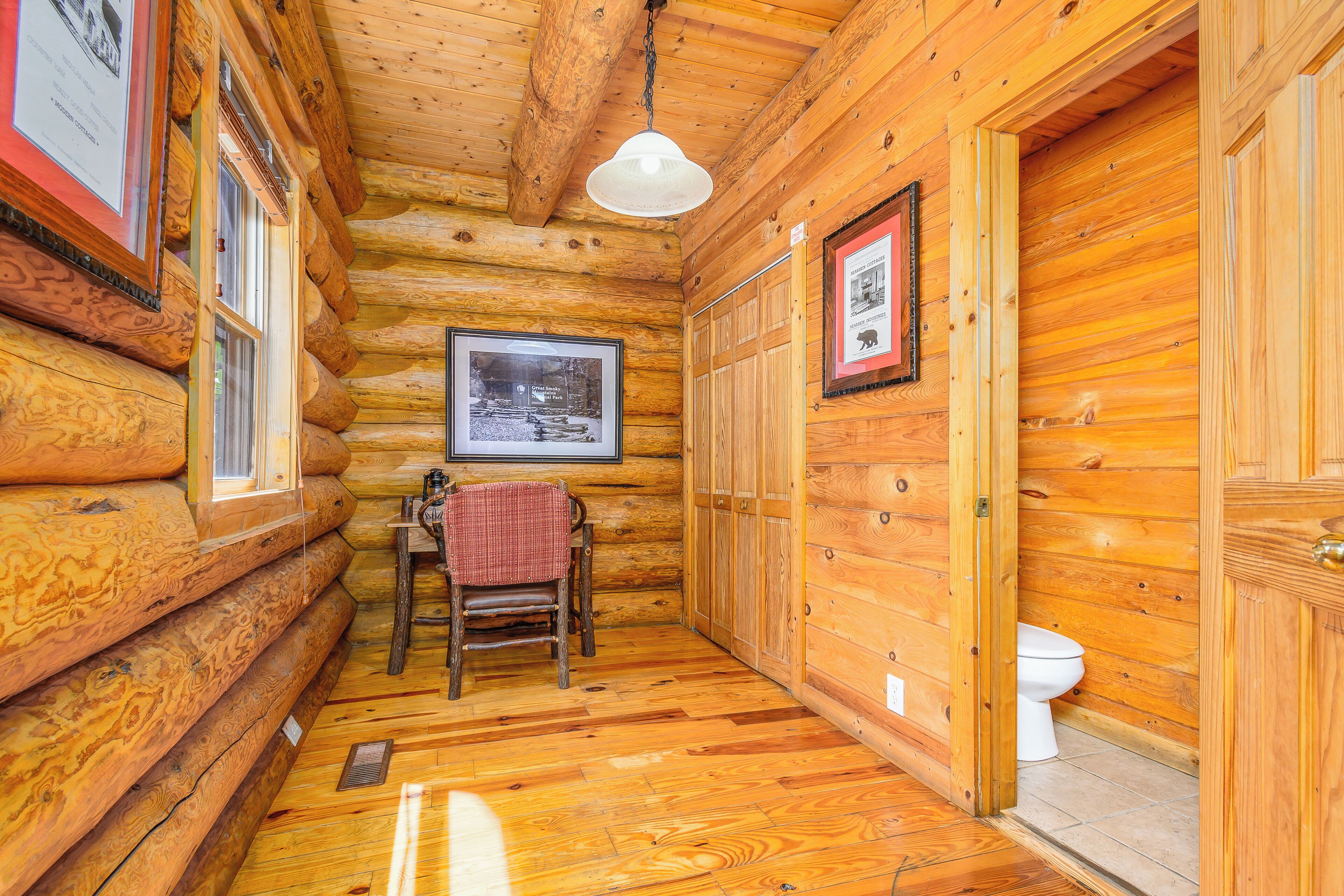 A log cabin bathroom with wooden walls, a wooden floor, a white toilet, a framed picture on the wall, and a hanging light fixture.