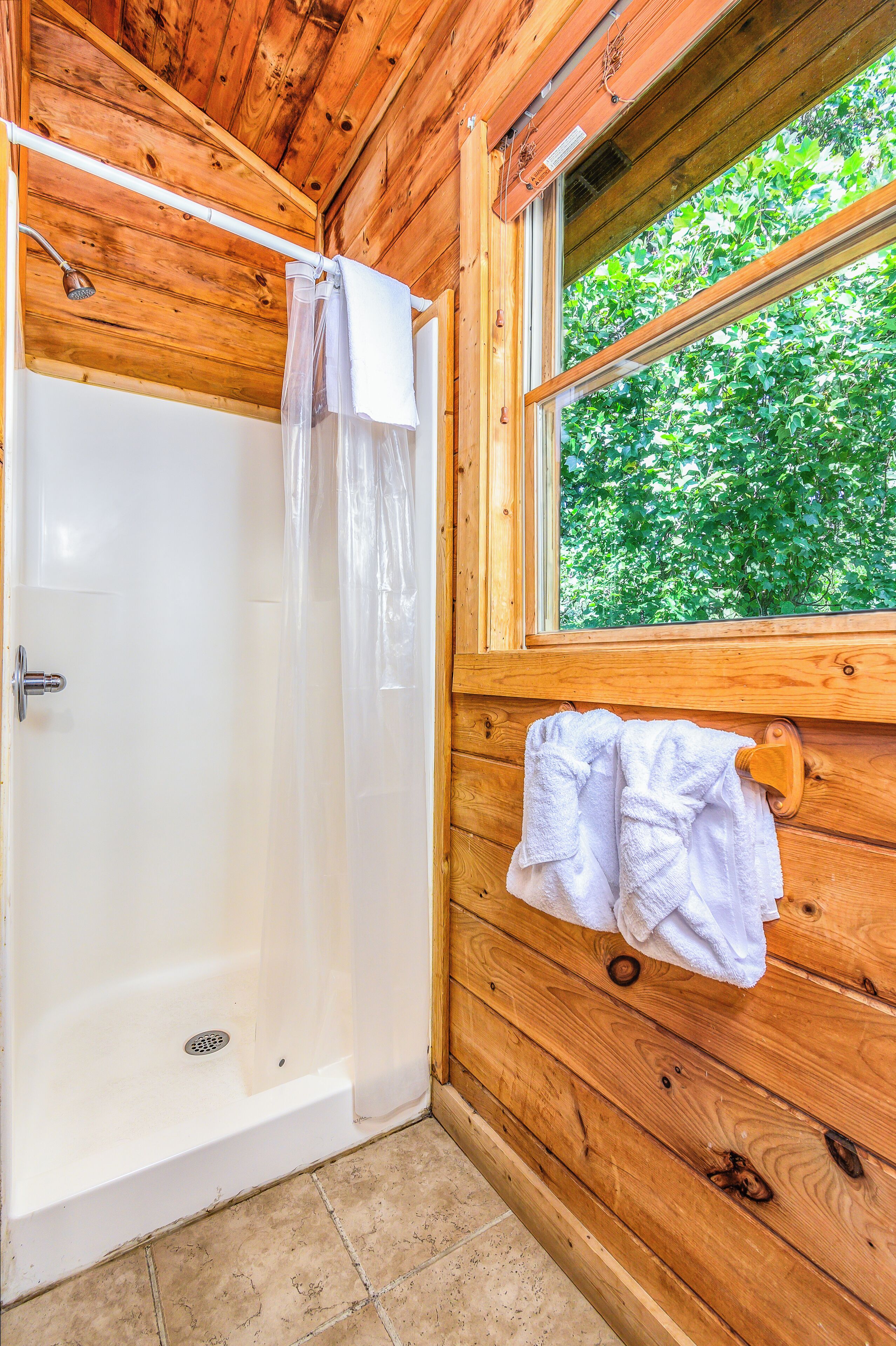 Un bagno in una cabina di legno con doccia, una finestra vista su del verde e un porta asciugamani con asciugamani.