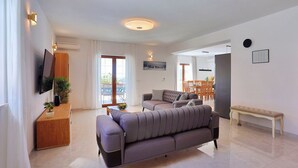 A modern living room with a grey sofa, a wooden side table, and a bench. There is a flat-screen TV mounted on the wall, and a dining area with a table and chairs is visible in the background. - Villa Nedy - Four Bedroom Villa, Sleeps 8 (Zakucac)