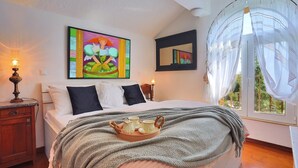 Ein Schlafzimmer mit einem Bett, einem Nachttisch, einer Lampe, einem Gemälde, einem Spiegel und einem Fenster mit durchsichtigen Vorhängen.