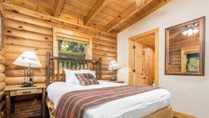A log cabin bedroom with a wooden bed, two bedside tables with lamps, a window, and a door.
