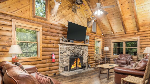A log cabin interior with a stone fireplace, leather furniture, and a mounted television.