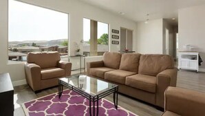 A spacious living room with a large window, a beige sofa, a glass coffee table, and a purple rug. - Amazing VIEWS, NEW quiet home, bonus Fitness Room! (Hurricane)