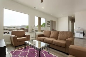 A spacious living room with a large window, a beige sofa, a glass coffee table, and a purple rug.