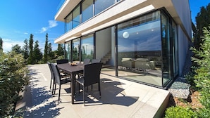 Una casa moderna con un patio que cuenta con una mesa y sillas, amplios ventanales y vistas del paisaje circundante.