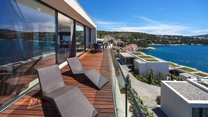 Een modern balkon met houten vloer, glazen leuning en lounge fauteuils, met uitzicht op een kustgebied met gebouwen en water.