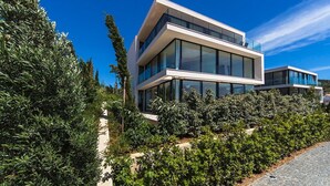 Een modern huis met twee verdiepingen, grote glasramen, omgeven door weelderige begroeiing en een helderblauwe lucht.