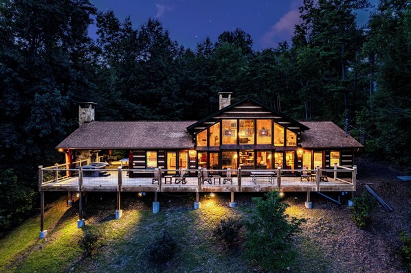 A two-story log cabin with a deck, surrounded by trees.
