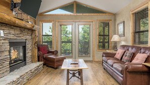 Ein gemütliches Wohnzimmer mit Kamin aus Stein, Ledersofas, einem Holztisch und großen Fenstern mit Jalousien.