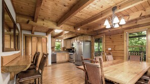 Interior rústico de una cabaña con pisos de madera, una mesa de comedor, sillas, una zona de cocina y un ventanal grande.